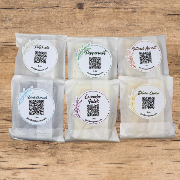 6 -1 oz bars of soap set out in 2 lines , all wrapped in white packaging with a  garden of green sticker that has a QR code and what kind of soap it is like patchouli, peppermint, oatmeal apricot, black charcoal, lavender fields, Italian lemon on each one. they are on a light wood background