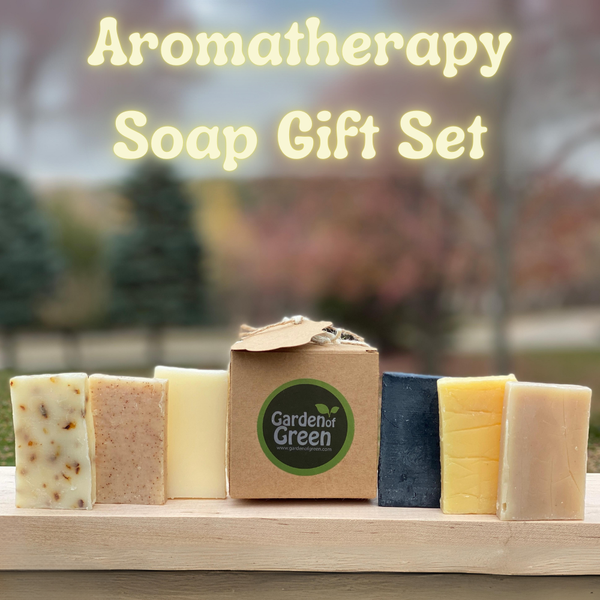 a big title on top of the picture that says "aromatherapy soap gift set" on top of the page in a light yellow. a kraft soap box with a garden of green sticker sits on a wood board with 3 different soaps on the left and 3 different soaps on the right. the background is pine trees and nature
