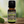 Cassia Garden of green essential oil front view sitting on a wood table with trees and leaves in the background. a stick of cinnamon on the table to the right of the bottle.