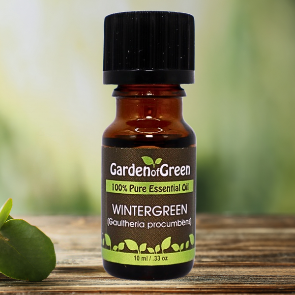 Wintergreen Garden of green essential oil front view sitting on a wood table with a leaf on the lower left hand side. green background with sun shining through the top left.