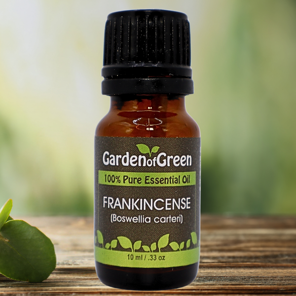 Frankincense Garden of green essential oil front view sitting on a wood table with a leaf on the lower left hand side. green background with sun shining through the top left.