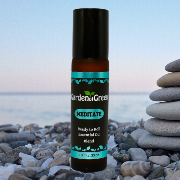 Meditate essential oil blend roller 10ml front view sitting on a bed of rocks and sea shells. on the right side there is a stack of smooth rocks. there is a light blue lake and sky in the background