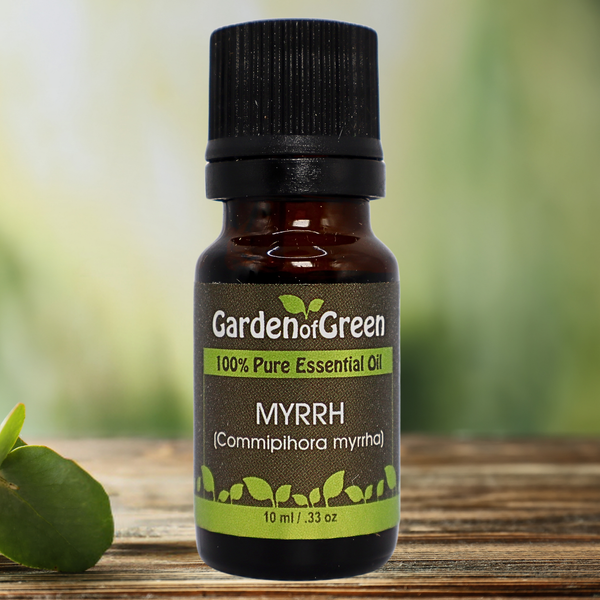 Myrrh Garden of green essential oil front view sitting on a wood table with a leaf on the lower left hand side. green background with sun shining through the top left.