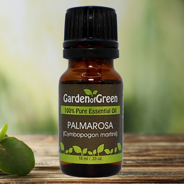 Palmarosa Garden of green essential oil front view sitting on a wood table with a leaf on the lower left hand side. green background with sun shining through the top left.