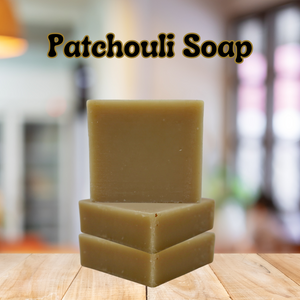 the title "patchouli Soap" is on top with black lettering with a tan outline. 3 bars of plain soap stacked on top of a wood table with a kitchen in the background